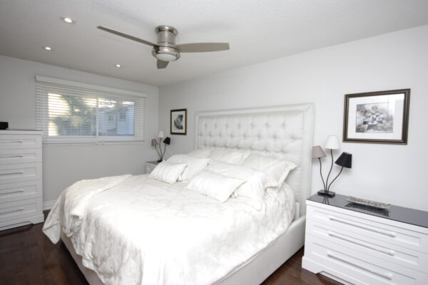 White bedroom with side tables and lamps on top and a ceiling fan and a window and paintings on the wall