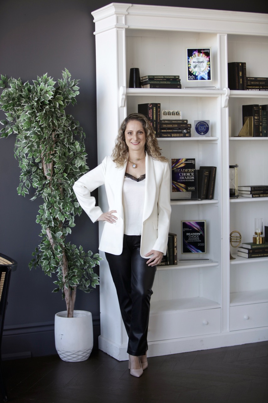 Woman in black and white standing against a bookshelf with awards on it and a tree plant in a pot beside