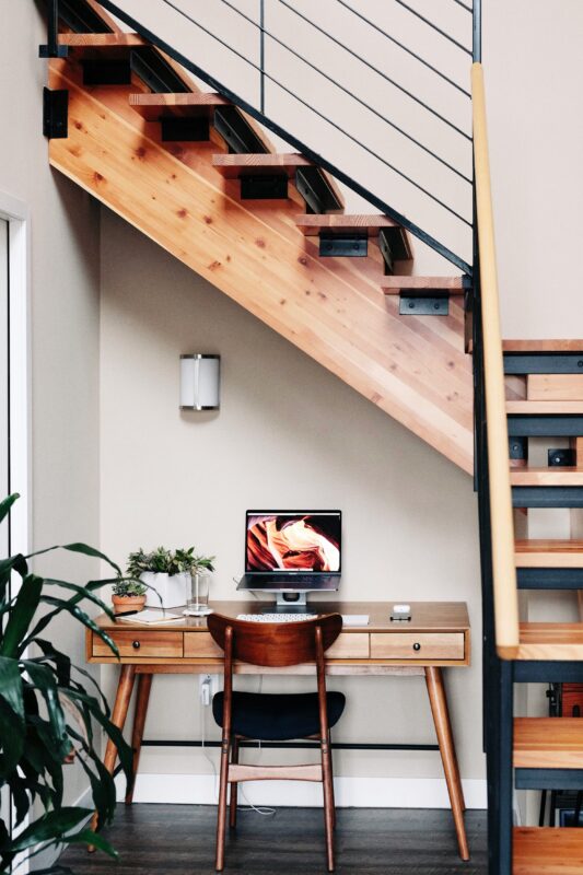 Minimalist home workspace under staircase with wooden desk and computer setup.